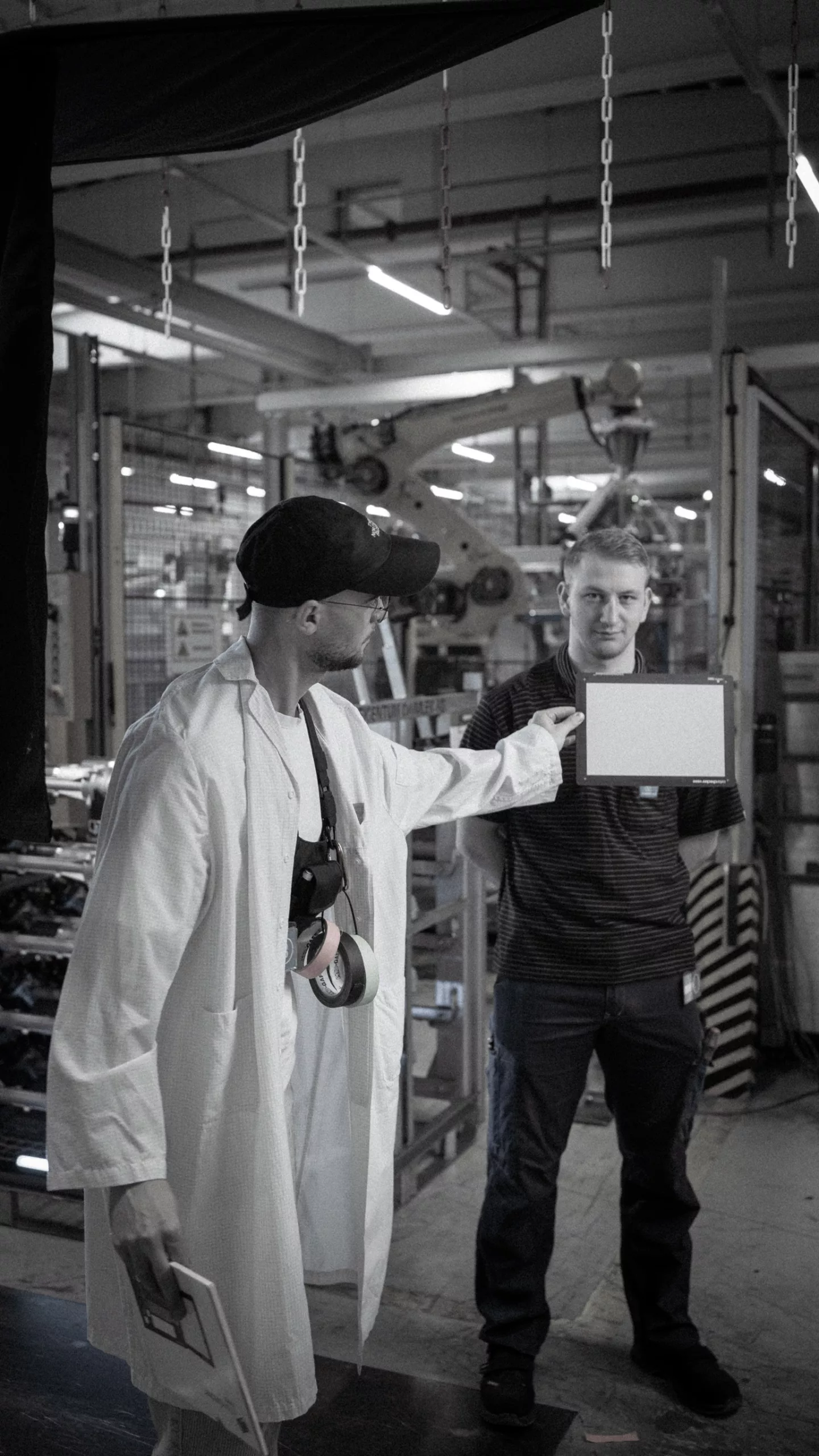 Produktion eines Werbevideos in einer Industriehalle – ein Videograf in weißem Kittel hält eine Farbkarte vor die Kamera, während ein Arbeiter im Hintergrund posiert